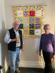 Two people standing in front of a hanging quilt 